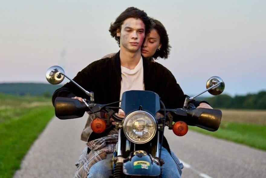 Still from a movie of two young people on a motorbike riding along an empty stretch of road, with green fields on either side of them.
