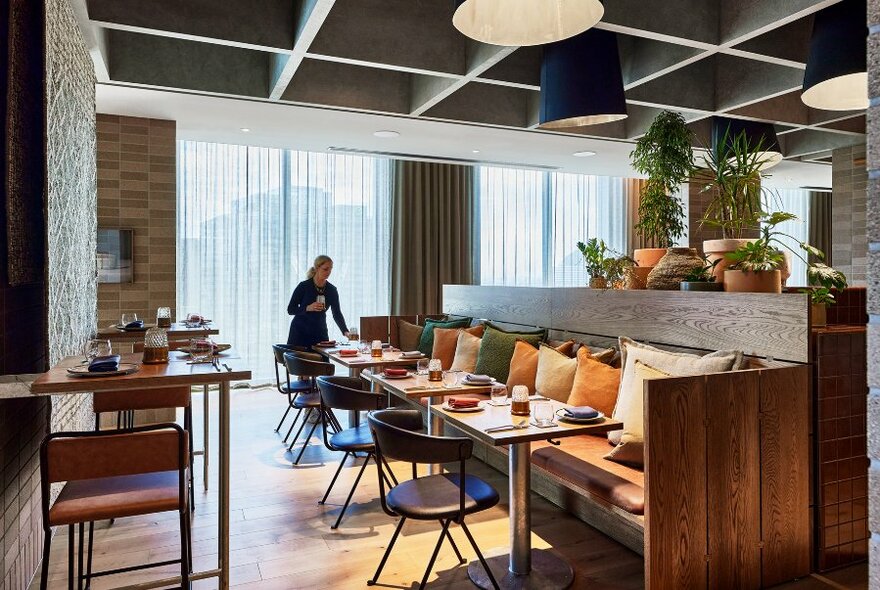 Waiter setting tables in restaurant space - high tables and stools to left, long wooden booth to right.