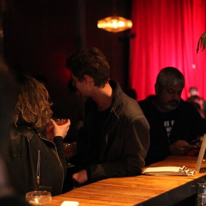 Two people leaning against a wooden bar, sipping drinks, soft lights and a red curtain in the background.