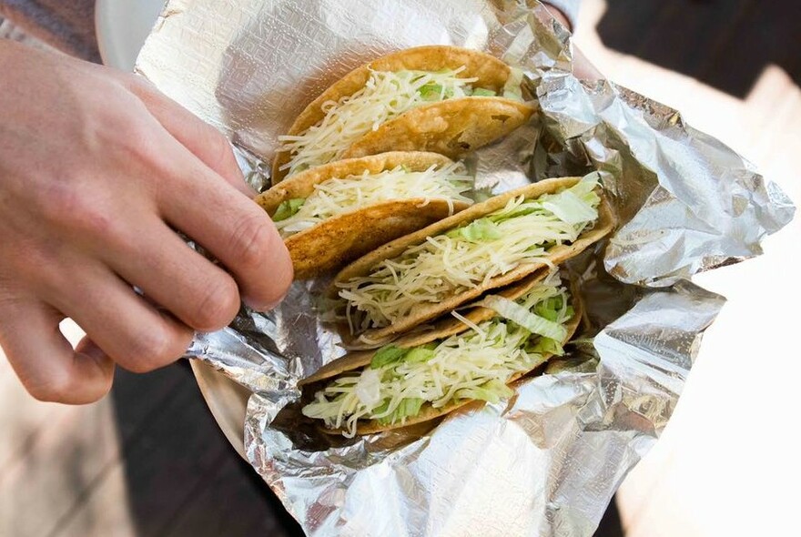 A hand reaching for one of four tacos in a foil wrapper.