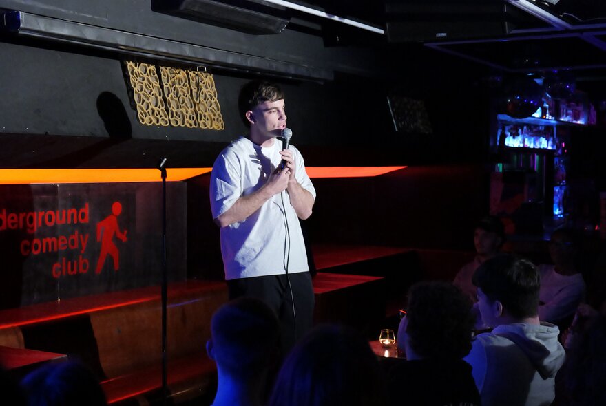 A live stand-up comedy performer in a dimly lit basement venue or club with an audience seated in the foreground.