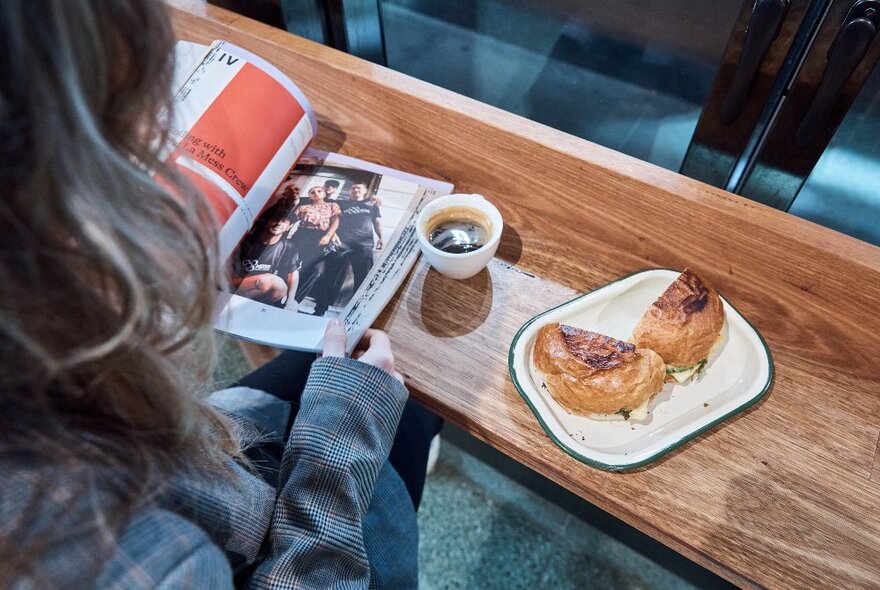 A woman, seen from behind, sitting at a narrow timber bench reading a magazine, with a gourmet sandwich and an espresso.