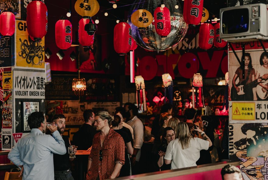 A vibrant, Japanese-inspired bar interior, featuring red hanging lanterns, postes on the walls and a lively crowd.