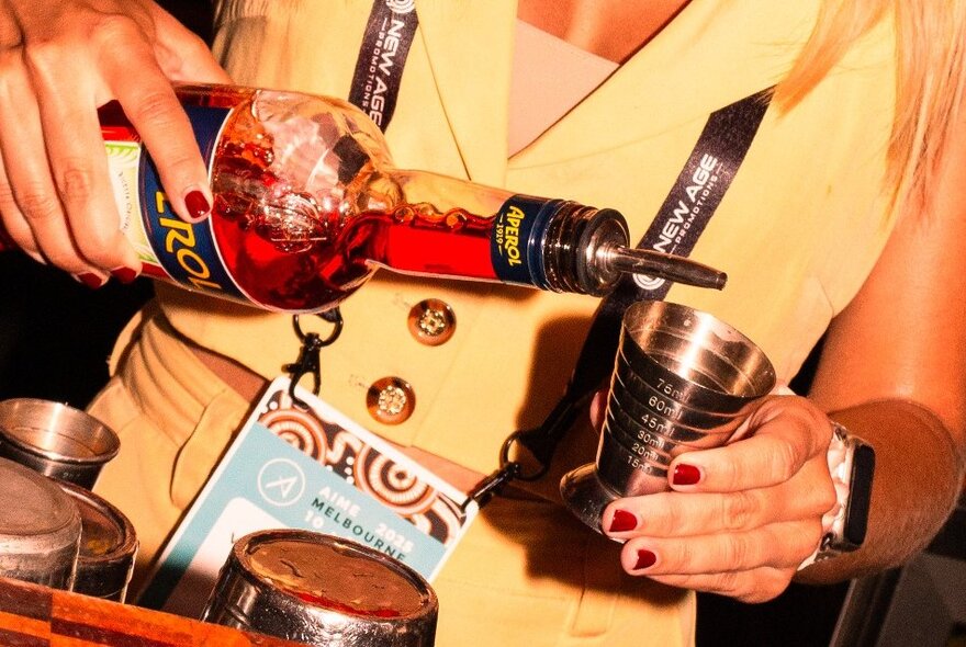 A person pouring out a measure of Aperol into a metal measuring cup.