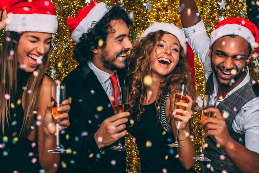 People wearing Santa hats laughing while holding champagne flutes, under a shower of sparkles.