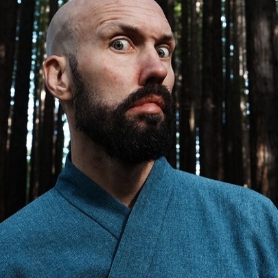 A close-up of a bald man with a prominent beard making a wide-eyed, comical expression. He is wearing a blue, textured robe-like garment.