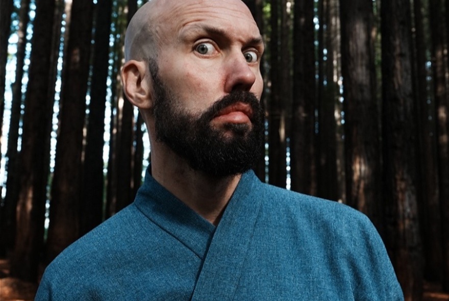 A close-up of a bald man with a prominent beard making a wide-eyed, comical expression. He is wearing a blue, textured robe-like garment.