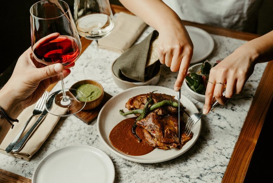 Looking down at a table in a restaurant, with hands cutting into a steak on a plate, and another hand holding a glass of red wine.