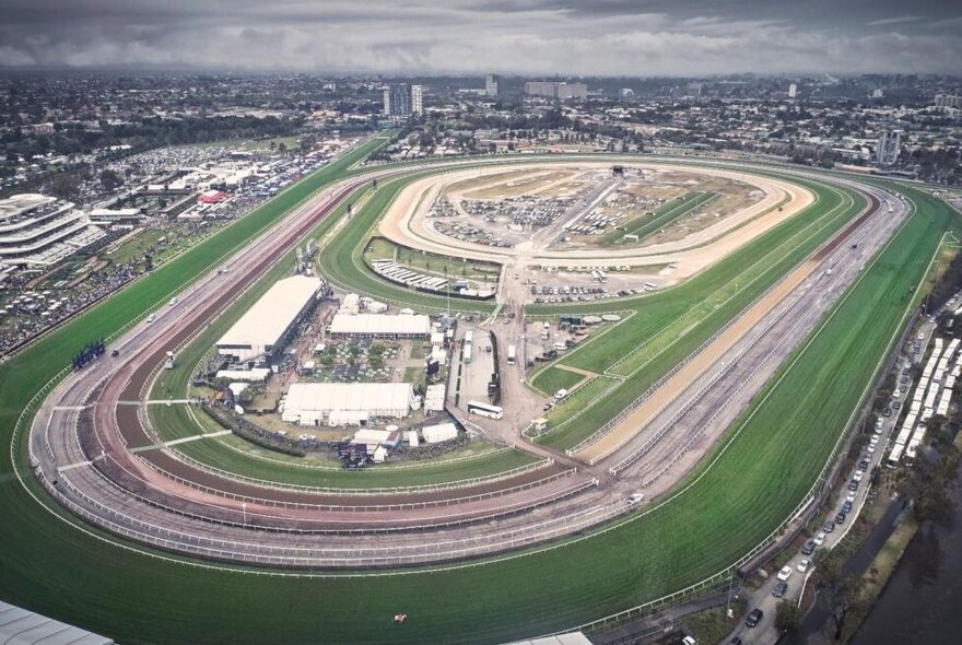Aerial view of Flemington Racecourse, a large, oval-shaped horse racing track with multiple concentric turf and dirt tracks, surrounded by green lawns and stadium structures under an overcast sky.