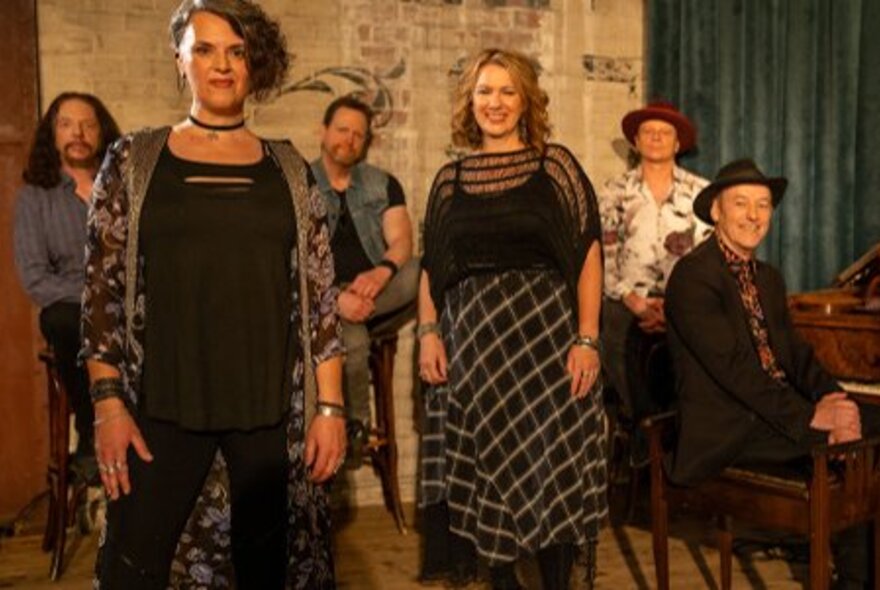 A group of musicians and singers, standing in a room with a piano, a green curtain behind them.
