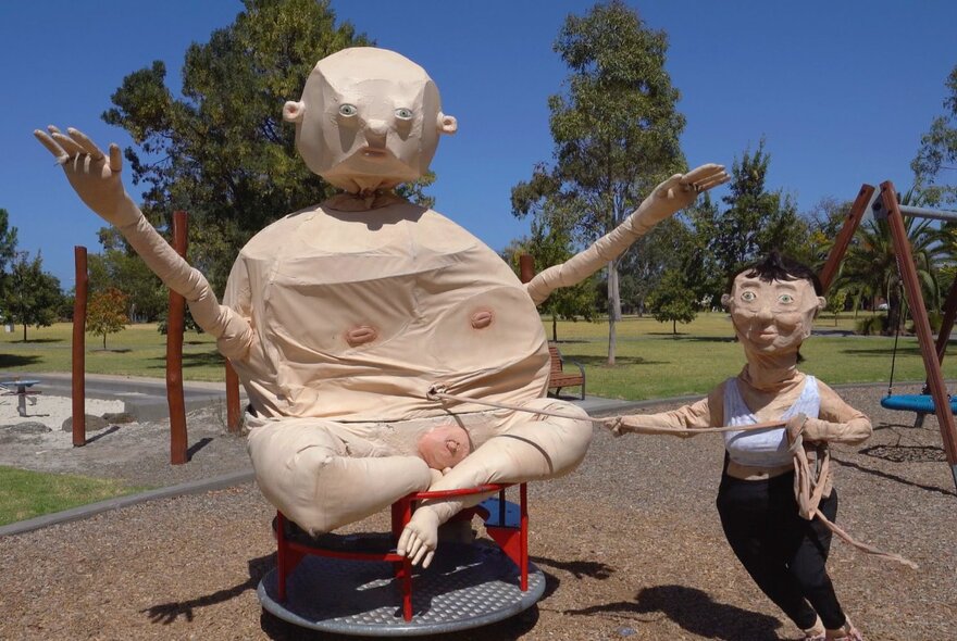 Woman wearing a mask holding a rope tied to a large puppet sculpture of a naked baby.