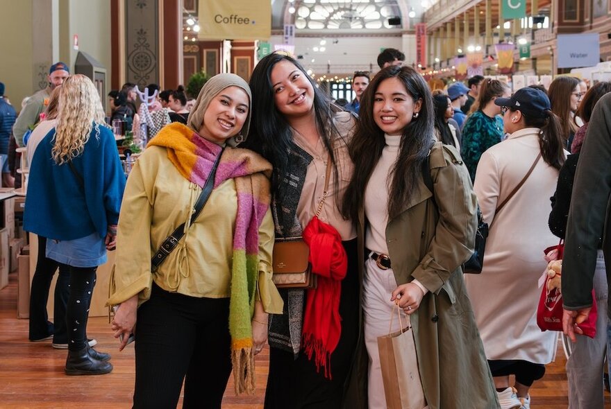 Three friends posed together smiling at an indoor design market stall, crowds of people wandering around the exhibition hall behind them.