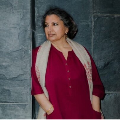 Author Geetanjali Shree, wearing a dark red long sleeved top and leaning casually against a dark grey stone wall.