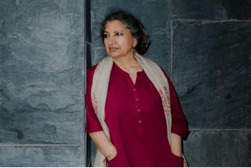 Author Geetanjali Shree, wearing a dark red long sleeved top and leaning casually against a dark grey stone wall.