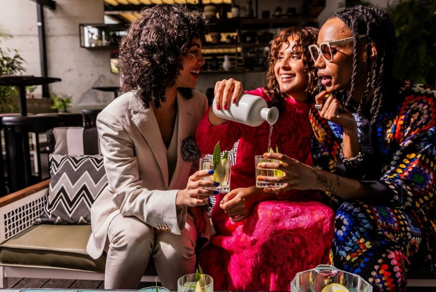 Three women seated on a bench in a bar environment with one woman pouring another a drink.
