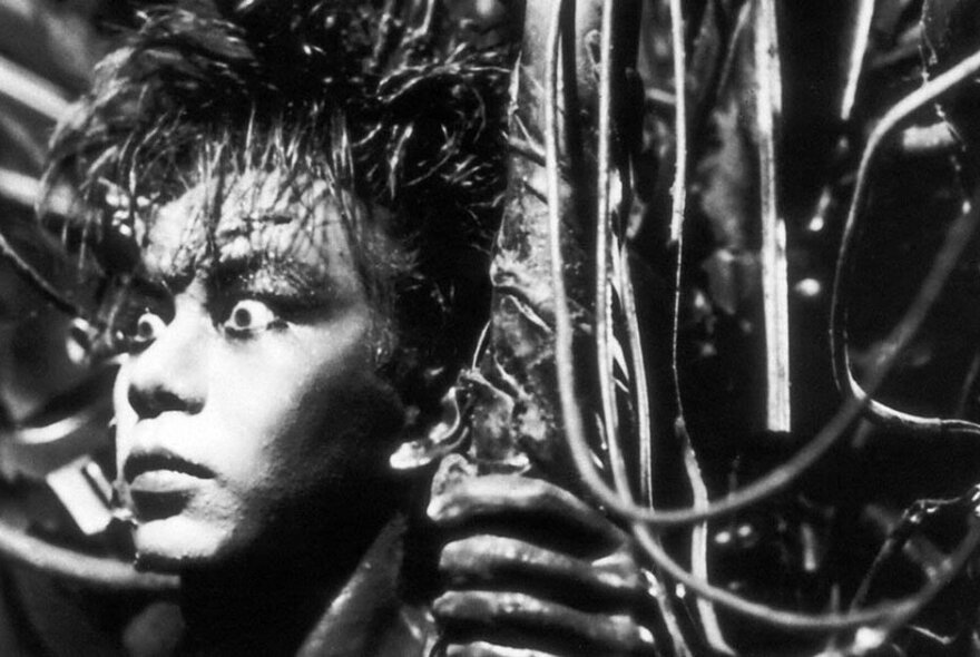 Black and white still from a Japanese horror film of a scary face with spikes in their forehead, their eyes bulging. 