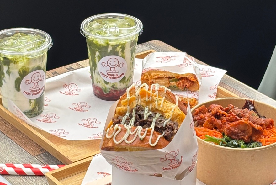 A Korean rice bowl, toasted sandwiches wrapped in paper, and two matcha drinks in plastic cups, on a table.