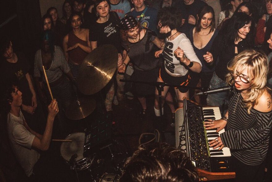 An intimate live music performance with a keyboard player and a drummer playing to an enthusiastic crowd in a dimly lit venue.