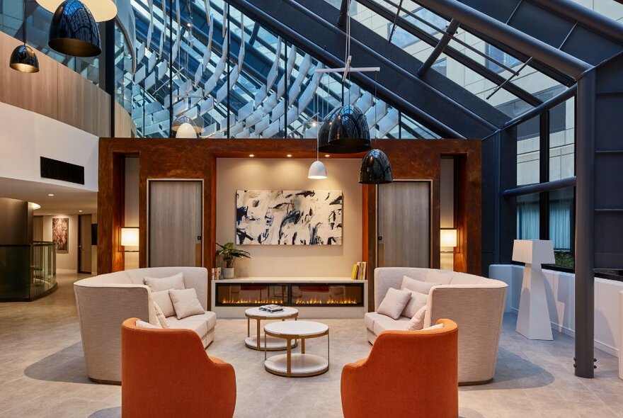 An empty hotel lobby with two orange tub chairs and two beige sofas under a glass ceiling. 