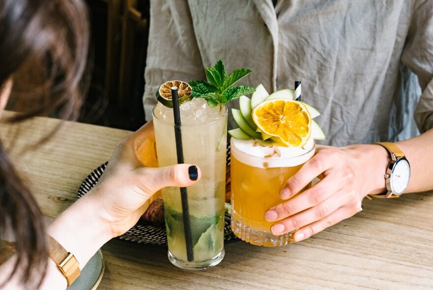 Two people clinking their cocktail glasses together over a timber table.