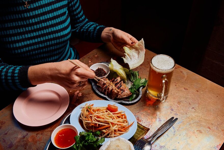 A woman in a blue and black striped top with chopsticks, eating from two Thai-style dishes presented on the table in front of her. 