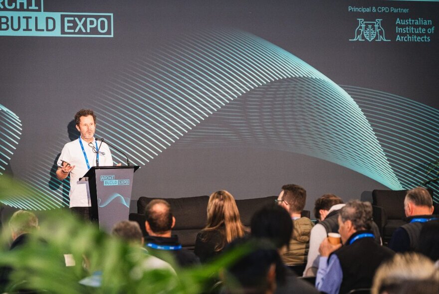 A speaker at a podium on stage in front of a seated audience, with a a large expo banner behind him.