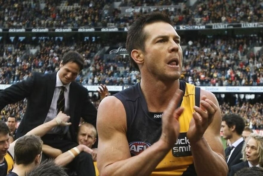 Former AFL star Ben Cousins clapping on the field in a packed stadium, while wearing the yellow and black jumper of the Richmond Football Club.