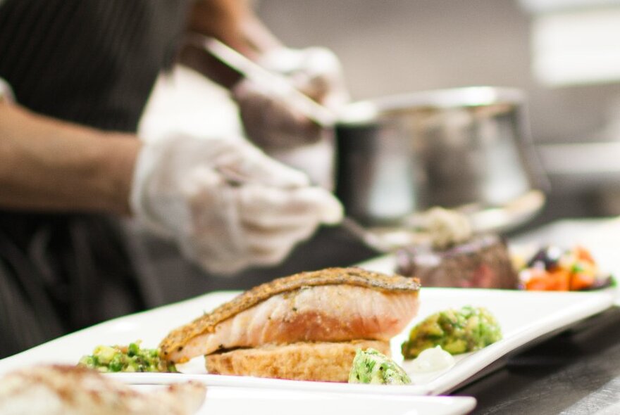Hands wearing white protective gloves holding a saucepan and adding ingredients to two plates of food on a counter.