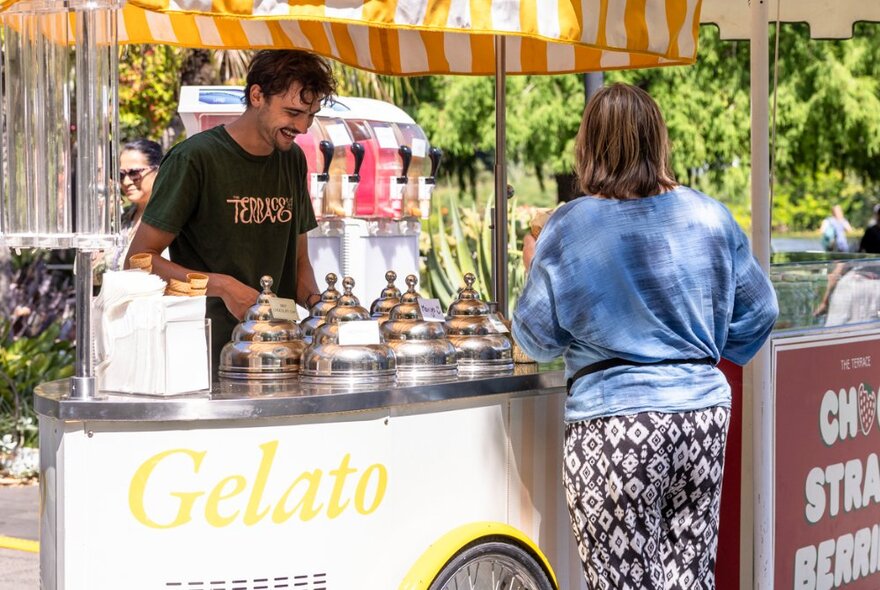The back of a woman at a gelato cart while a smiling man serves her. 