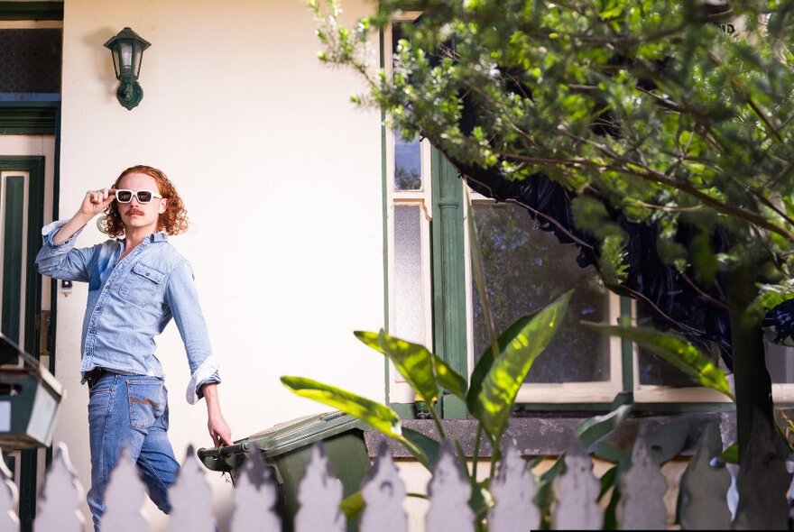 Comedian with curly red hair and glasses standing at the front door of a Victorian cottage.