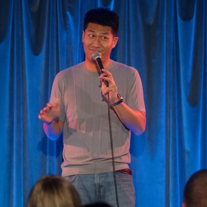 A comedian on a small stage in front of a blue curtain making a face into a microphone.