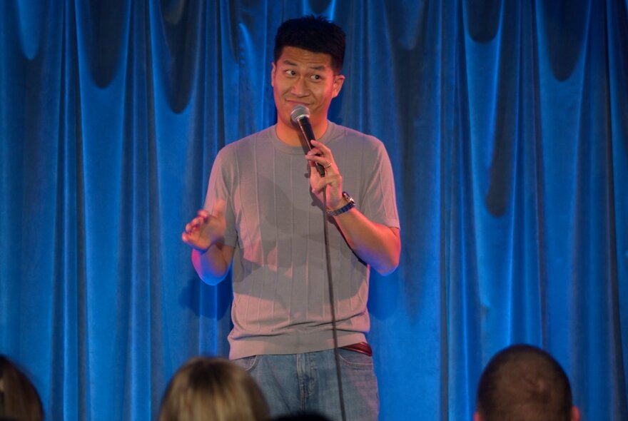 A comedian on a small stage in front of a blue curtain making a face into a microphone.