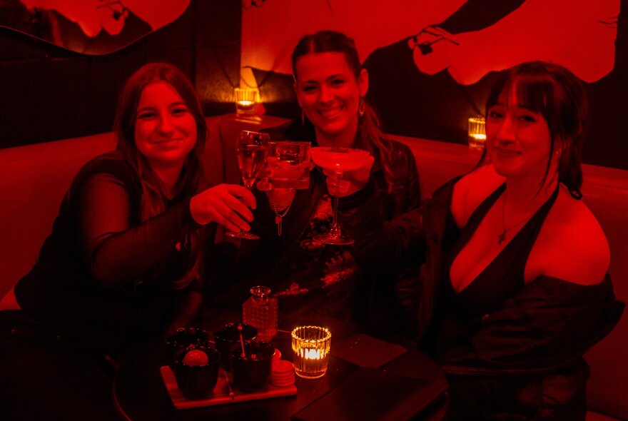 Three friends seated at a booth enjoying drinks, cast in a red light.