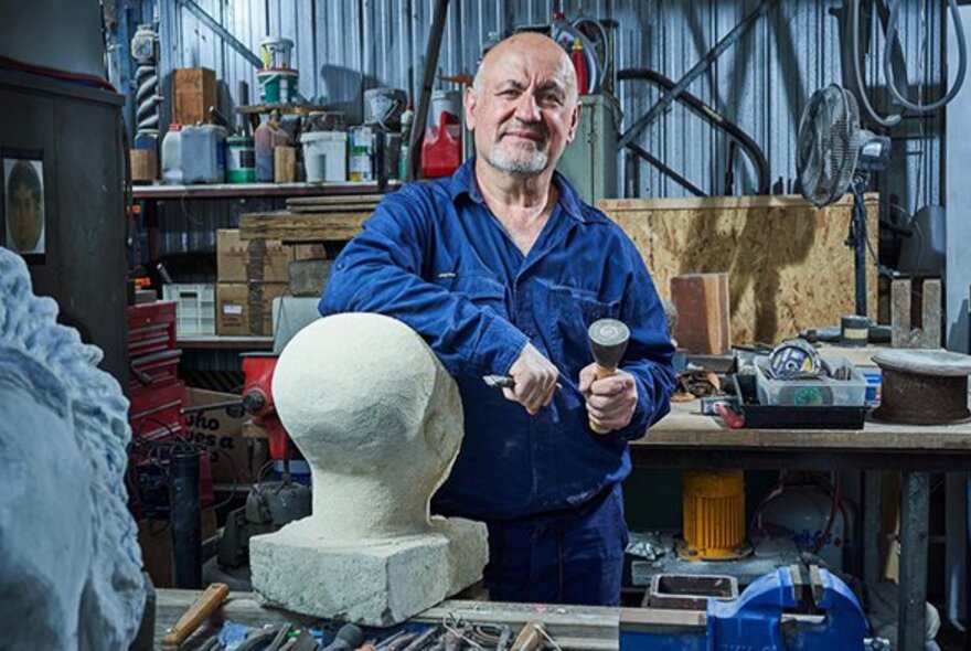 A man in a blue shirt standing behind a marble sculpture and holding sculpting tools in his hands, in a workshop.
