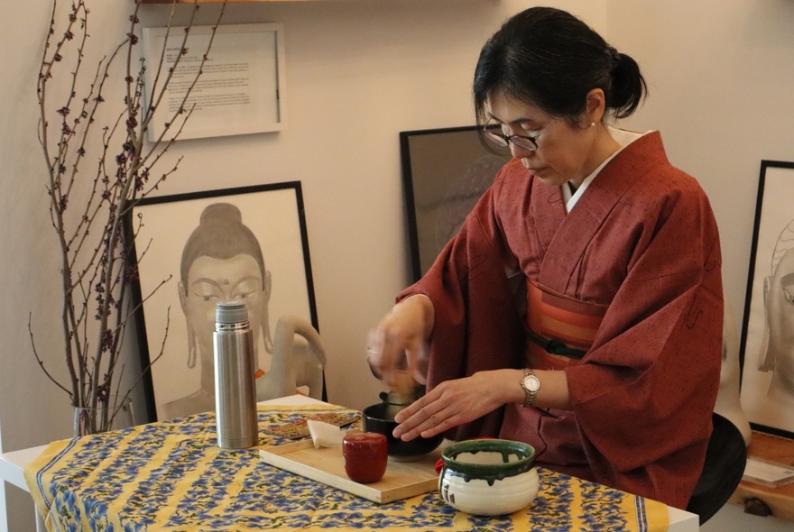 A Japanese woman dressed in a deep red kimono, at a small table with traditional Japanese Tea Ceremony implements in front of her.