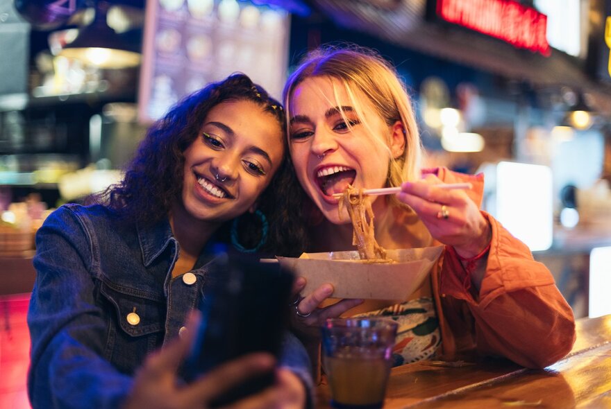 Two young women taking a selfie together while one of them eats a bowl of noodles.