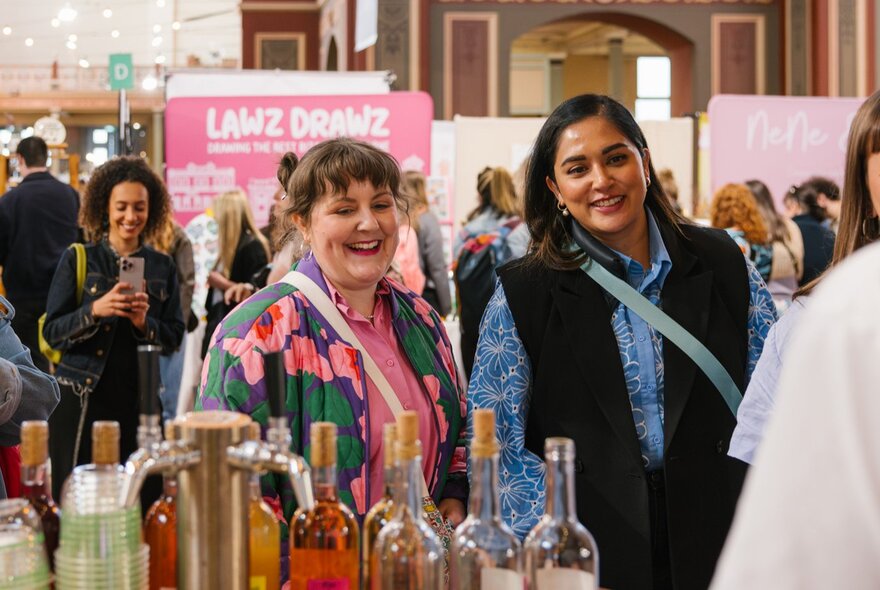 Two women laugh and interact at a market stall with many bottles of coloured drinks. 