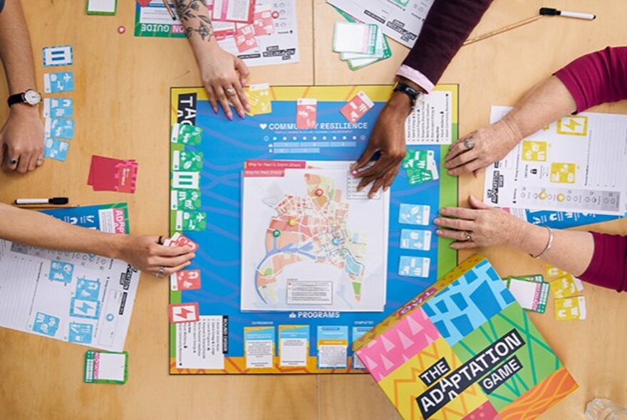 Top down view of hands leaning across a board game on a table and moving the tokens and cards, the group of people are in the act of playing the game.