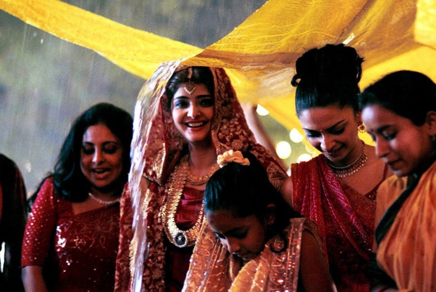 Women at an Indian wedding, wearing jeweled robes, standing under yellow cloth.