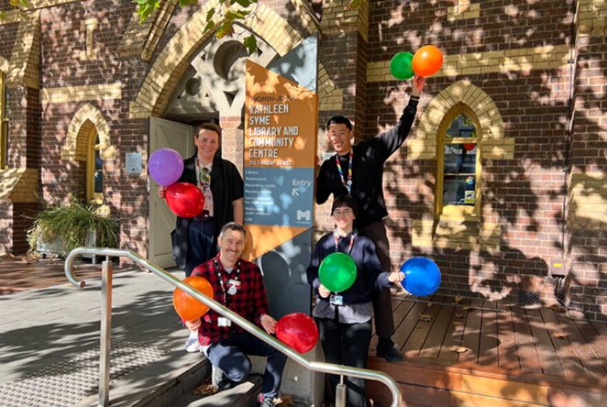Workers at the Kathleen Syme Library holding balloons at the entrance sign, the brick library behind them.
