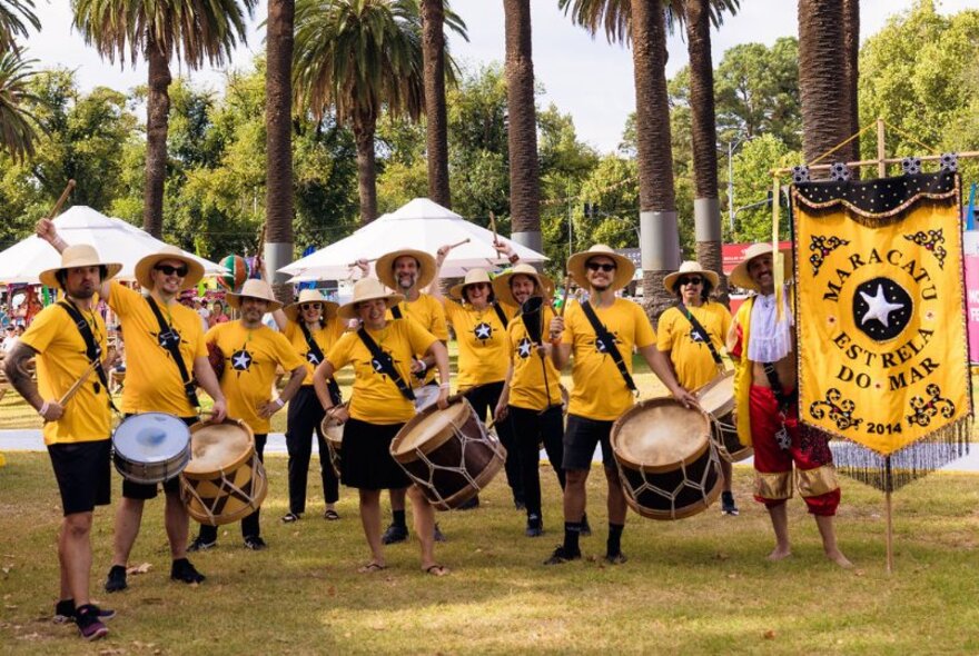 A group of people wearing yellow t-shirts and straw hats, playing Afro-Brazilian drums.