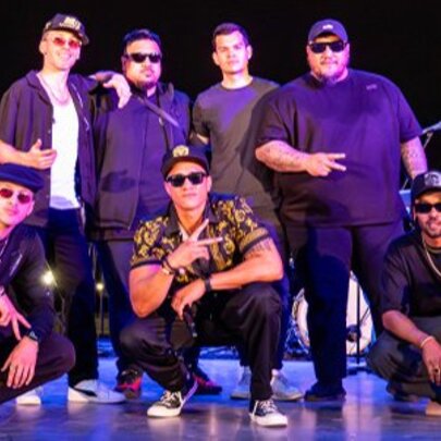 Seven men on stage, most wearing sunglasses and caps, posing like a boy band. 