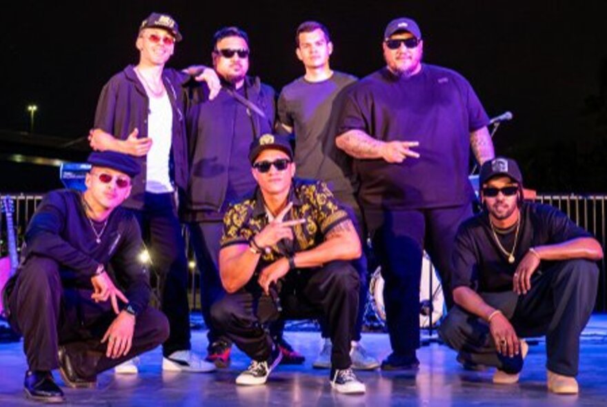 Seven men on stage, most wearing sunglasses and caps, posing like a boy band. 