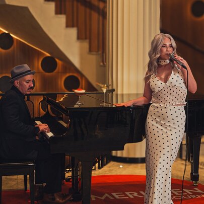 A man seated at a grand piano plays while a woman in a white spotted evening dress sings in a hotel lobby. 