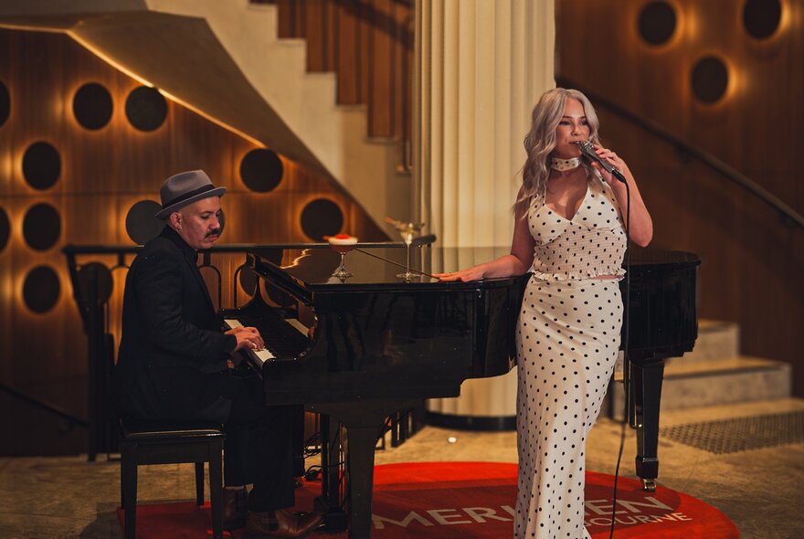 A man seated at a grand piano plays while a woman in a white spotted evening dress sings in a hotel lobby. 