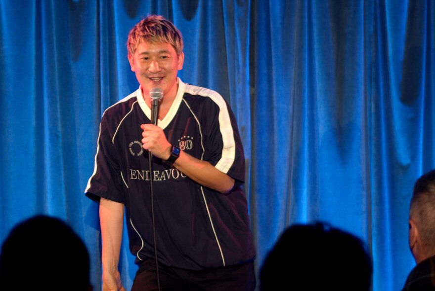A stand-up comedian talking into a microphone they hold in their hand, performing live on stage.