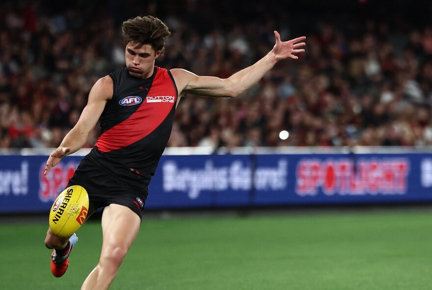 An Essendon AFL player running while about to kick a yellow football he has just dropped in front of his leg, with blurred fans in the stadium in the background.