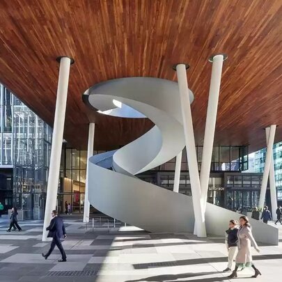 The modern outdoor plaza at Melbourne Quarter precinct, featuring a large, white spiral staircase as its centerpiece, situated under a massive wooden ceiling supported by angled white pillars.