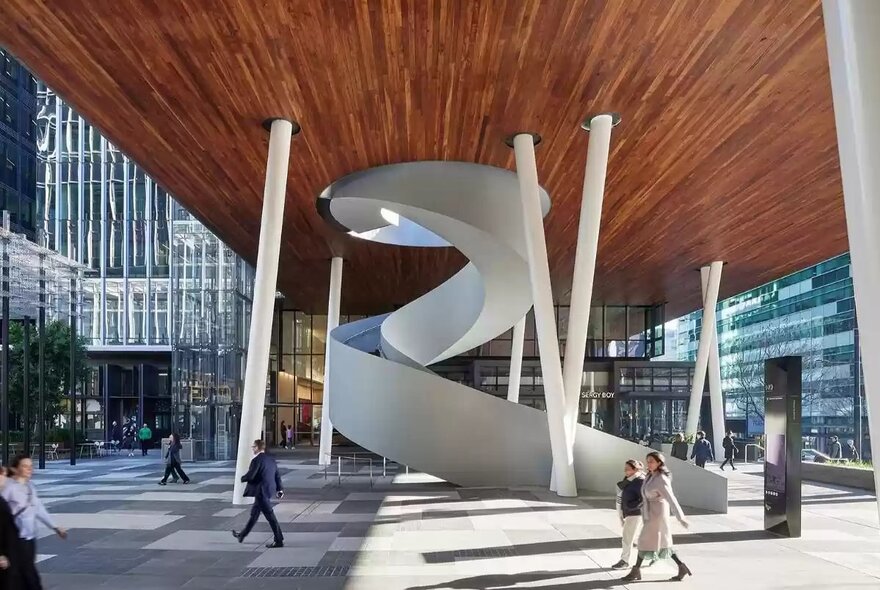 The modern outdoor plaza at Melbourne Quarter precinct, featuring a large, white spiral staircase as its centerpiece, situated under a massive wooden ceiling supported by angled white pillars.