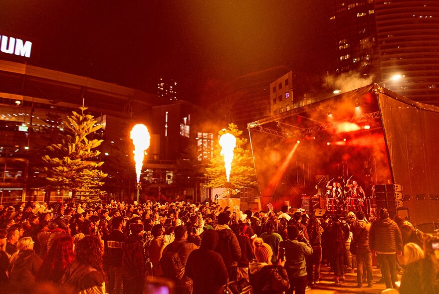 A crowd at the Firelight Festival with an orange glow and fire shooting into the air. 